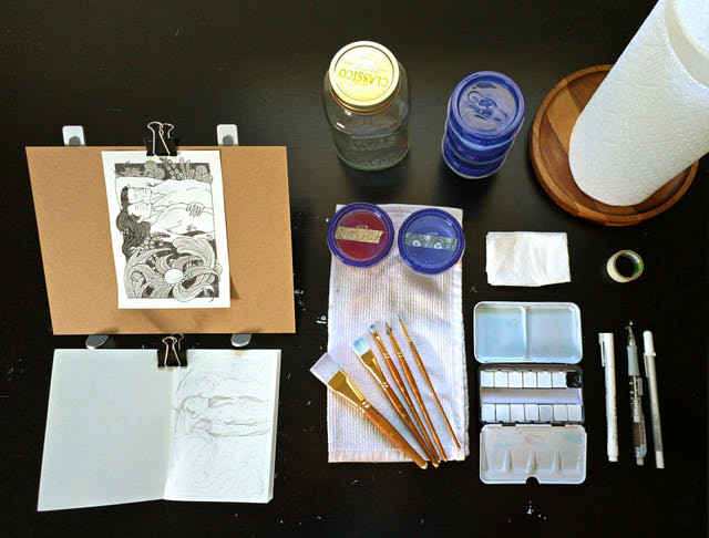 A spread of watercolor supplies, with a grayscale painting towards the left.